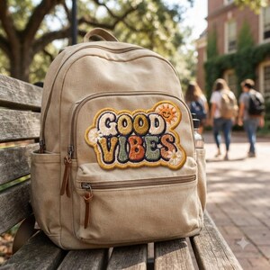 Könnte beinhalten: Ein beigefarbener Rucksack aus Segeltuch mit einem großen gestickten Aufnäher auf der Vordertasche. Der Aufnäher zeigt den Schriftzug "GOOD VIBES" in einer retro-bunten Schriftart, akzentuiert durch kleine Blumenstickereien und eine lachende Sonne.