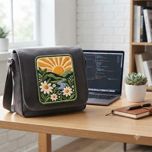 May include: A charcoal gray messenger bag featuring a rectangular embroidered patch on the front flap. The embroidery depicts a vibrant yellow sun rising over green rolling hills, a winding blue river, and white daisy flowers with green leaves.