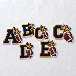 Puede incluir: Un conjunto de cinco parches bordados con las letras A, B, C, D y E en azul marino con bordes dorados. Cada letra está decorada con una corona dorada y un gráfico de balón de fútbol americano marrón con cordones blancos sobre tela blanca.