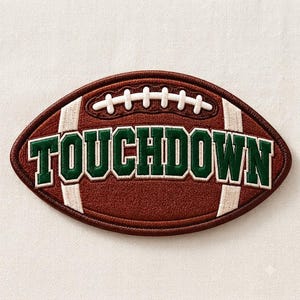 Puede incluir: Un parche bordado de forma ovalada diseñado como un balón de fútbol americano marrón. El parche presenta cordones blancos en la parte superior y la palabra TOUCHDOWN bordada en letras verdes negritas con un borde blanco en el centro.