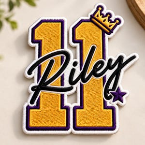 Puede incluir: Un parche bordado que muestra el número 11 en amarillo con un borde morado. El nombre Riley está cosido en letra cursiva negra sobre los números. Una pequeña corona amarilla se sitúa sobre el segundo número y una estrella morada está abajo a la derecha.