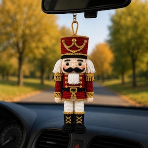 Può includere: Un ornamento a forma di schiaccianoci all'uncinetto appeso a uno specchietto retrovisore. La figura presenta una giacca e un cappello rossi con finiture dorate, baffi neri, capelli e barba in filato bianco, pantaloni bianchi e stivali neri.