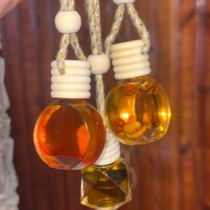 May include: Three small glass hanging diffuser bottles filled with amber and gold-toned liquid. Two bottles are spherical and one is faceted, each topped with a ribbed wooden cap and suspended by a thick, tan, metallic-flecked cord with wooden beads.