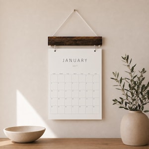May include: A wall-hanging calendar for January 2027 featuring a white paper sheet with black text and a grid layout. The calendar is suspended from a dark brown wooden bar with a white cord, providing a clean and functional display for the month.
