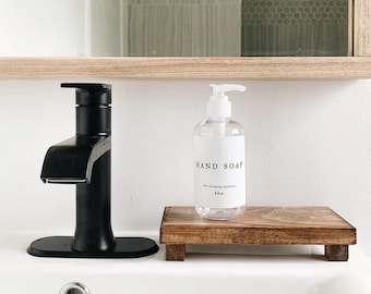 May include: A clear plastic hand soap bottle with a white pump and a label reading HAND SOAP by:ninasprojects 8fl oz, resting on a small rectangular wooden riser next to a matte black bathroom faucet on a white sink basin.