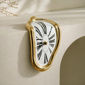 May include: A gold-toned, melting-style wall clock with a white face and black Roman numerals. The clock features a distorted, asymmetrical frame that curves over the edge of a surface, with black hour and minute hands at the center.