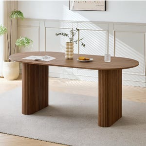 Pode incluir: Uma mesa de jantar oval de madeira com uma superfície castanha lisa e duas bases de pedestal cilíndricas, grossas e caneladas. A mesa está disposta com um livro aberto, um vaso texturizado de cor creme com caules verdes, um prato pequeno com comida e um copo.