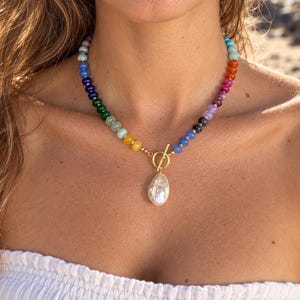 May include: A necklace featuring a strand of multicolored round beads in a rainbow gradient, connected by a gold-toned toggle clasp. A single large, irregular white baroque pearl pendant hangs from the center of the toggle closure.