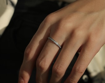 May include: A thin, silver-toned band ring with a textured, glittery surface finish. The ring features a slim, uniform profile that encircles the finger, reflecting light across its granular, metallic exterior texture for a subtle sparkle.