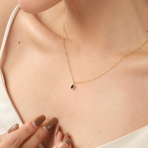 Peut inclure: Un collier à fine chaîne dorée doté d'un petit pendentif rectangulaire orné d'une pierre noire lisse et polie en son centre. Le collier repose sur la clavicule, présentant un design épuré avec une fine chaîne à maillons métalliques brillants.