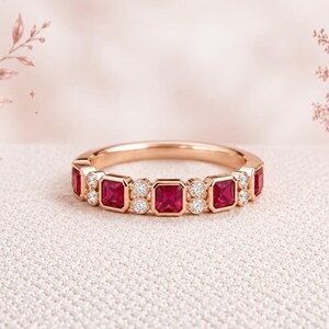 May include: A rose gold band ring featuring an alternating pattern of rectangular-cut red rubies and small round white diamonds. The gemstones are set in a bezel style along the top half of the metal band, creating a continuous, symmetrical design.