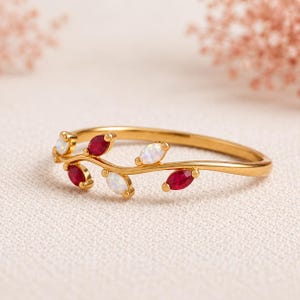 Peut inclure: Une bague en or dotée d'un anneau délicat en forme de vigne, orné d'une alternance de rubis rouges et d'opales blanches irisées taillés en marquise. L'anneau métallique se courbe doucement, imitant la forme organique d'une branche.