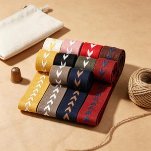 May include: A collection of woven fabric ribbons in various colors including yellow, beige, navy blue, red, brown, black, pink, and cream, each featuring a repeating chevron pattern. A metal thimble and a ball of twine are positioned nearby.