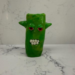 May include: A cylindrical green sculpture with a glossy finish, featuring two pointed ears on the sides, two round eyes with red pupils, and a mouth displaying a row of small white teeth. The object has a smooth, molded texture throughout.