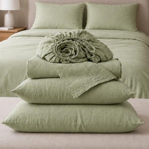 May include: A sage green bedding set featuring a fitted sheet, a flat sheet, and two pillowcases. The items are stacked neatly in the foreground, showcasing a textured fabric finish. The set matches the sage green duvet cover on the bed behind.