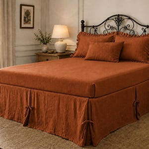 May include: A rust-colored bed set featuring a fitted sheet, two ruffled pillow shams, one rectangular throw pillow, and a bed skirt with tie-side details. The bedding is made of a textured fabric and is displayed on a bed with a metal frame.