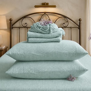 May include: A set of light mint green bedding featuring two stacked rectangular pillows, a folded flat sheet, and a fitted sheet with a textured, crinkled fabric finish. The bedding is displayed on a bed surface with a matching light mint green cover.