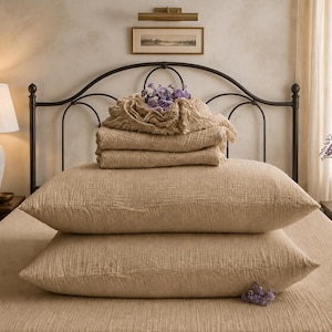 May include: A set of beige bedding featuring two rectangular pillows stacked horizontally, topped with two folded flat sheets and a gathered fabric piece. The textile has a visible crinkled, textured surface throughout the entire bedding set.
