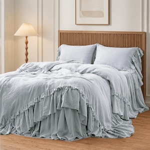 May include: A light blue bedding set featuring a duvet cover and two pillowcases with ruffled edges. The fabric has a soft, textured appearance, and the duvet cover drapes over the sides of the bed with a layered, gathered ruffle trim detail.