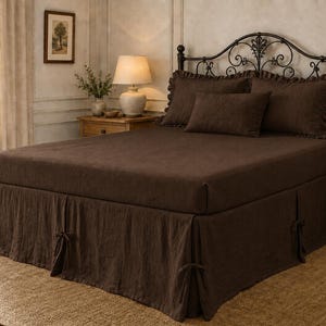 May include: A dark brown bed ensemble featuring a fitted sheet, a flat sheet, and multiple pillows with ruffled edges. The bed includes a matching bed skirt with decorative tie closures at the corners and sides, set against a black metal headboard.