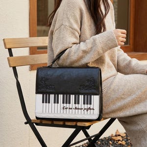 May include: A black and white rectangular handbag featuring a piano keyboard graphic on the front flap, ornate embossed corner details, and the cursive text C'est ma chanson préférée printed in brown ink above a white lace trim border.