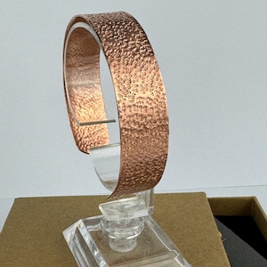 Pode incluir: Uma pulseira larga de cobre com uma superfície texturizada e martelada no exterior. O metal tem um acabamento metálico quente de cor castanho-avermelhada com um padrão repetitivo de covinhas em toda a banda exterior, com aspeto polido.