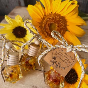 May include: Three small, faceted glass bottles filled with amber liquid and dried petals, each topped with a light wood cap and tied with a metallic gold and white cord. A brown paper tag on one bottle reads 8ML CAR DIFFUSER GOLDEN HOUR SUNFLOWER.