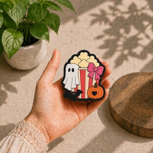 Puede incluir: Una pegatina troquelada que muestra un cubo de palomitas de rayas rojas y blancas lleno de palomitas, decorado con un fantasma blanco, un murciélago negro, un lazo rosa brillante y una calabaza naranja con un corazón negro en la parte frontal.