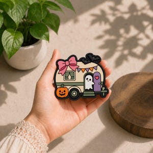 May include: A decorative rubber patch shaped like a vintage camper trailer. The cream-colored camper features a large pink bow, a smiling ghost in the doorway, a jack-o'-lantern, orange and purple bunting, and two black bats on the roof.
