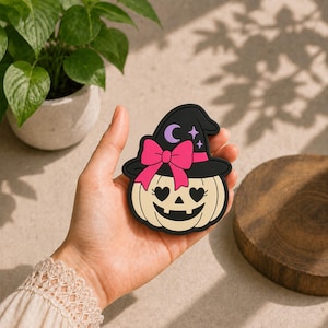 Puede incluir: Un parche decorativo plano con forma de calabaza color crema que lleva un sombrero de bruja negro. El sombrero presenta un lazo rosa brillante, una luna creciente morada y pequeñas estrellas moradas. La calabaza tiene ojos en forma de corazón.