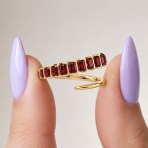 May include: A gold bypass ring featuring a row of ten rectangular, deep red garnet gemstones set in a channel. The ring has a polished gold band that wraps around, with the gemstone-set portion overlapping the plain gold section of the band.
