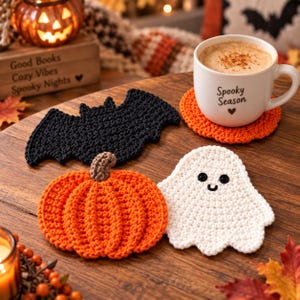 May include: A set of three crocheted coasters featuring a black bat, an orange pumpkin with a brown stem, and a white ghost with black eyes and a smile. A white ceramic mug with the text Spooky Season and a heart sits on an orange coaster.