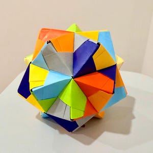 Puede incluir: Una esfera de origami kusudama multicolor construida a partir de unidades de papel plegado. La estructura geométrica presenta un patrón repetitivo de facetas triangulares y pentagonales en tonos vibrantes de naranja, amarillo, azul, azul claro y verde lima.