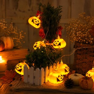May include: A string of orange LED jack-o'-lantern lights featuring carved facial expressions. The glowing pumpkin-shaped bulbs are connected by a thin wire and are arranged around a small green plant in a white picket fence planter.