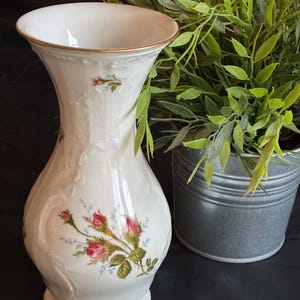 Könnte beinhalten: Eine weiße Porzellanvase mit einem zarten geprägten Rankenmuster und einem bemalten Blumenmotiv aus rosa Rosenknospen mit grünen Blättern. Die Vase hat einen ausgestellten Rand und einen Fuß, die beide mit einem dünnen Goldrand verziert sind.