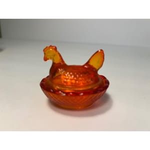 May include: A small, translucent orange glass hen on a nest dish. The lid features a moulded chicken figure with detailed feather textures, while the base is a round bowl with a cross-hatched diamond pattern around the exterior rim.