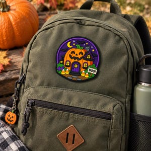 May include: An olive green canvas backpack featuring a circular patch with a carved pumpkin house design. The patch displays a smiling jack-o'-lantern structure with a purple door, yellow windows, candy corn, and a sign that reads BOO! A small pumpkin charm hangs from the zipper.
