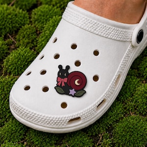 May include: A white foam clog shoe featuring a decorative charm shaped like a black snail with a dark red shell, a pink bow, a yellow crescent moon, and a light purple star, all resting on two green leaves attached to the shoe's ventilation holes.
