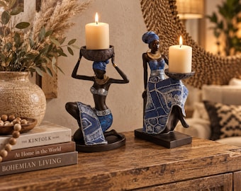 Puede incluir: Dos portavelas decorativos con figuras de piel oscura que visten ropa y pañuelos con patrones azules y blancos. Una figura se arrodilla mientras equilibra un cuenco para una vela en la cabeza, mientras la otra se sienta sosteniendo un plato.