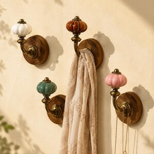 May include: Four wall-mounted hooks featuring round wooden bases and brass-toned metal arms. Each hook is topped with a fluted ceramic knob in white, brown, teal, or pink. A beige patterned scarf hangs from the brown knob, and a gold chain hangs from the pink knob.