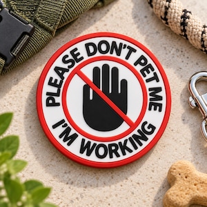 May include: A circular red, white, and black patch featuring a graphic of a black hand inside a red prohibition circle. The text reads PLEASE DON'T PET ME I'M WORKING in bold black capital letters arranged around the central hand icon.