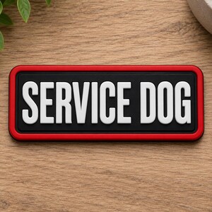 May include: A rectangular patch with a black background and a bold red border. The text SERVICE DOG is embossed in large, white, sans-serif capital letters across the center of the patch. The design features a clean, high-contrast aesthetic.