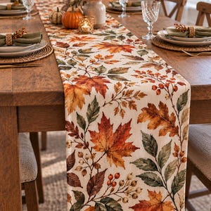 May include: A rectangular table runner featuring a printed pattern of autumn leaves in shades of orange, red, brown, and green, interspersed with clusters of small berries on a cream-colored fabric background, draped over a wooden dining table.