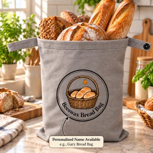 May include: A light gray fabric bread bag with two side handles and a snap closure. The front features a circular graphic of a woven basket filled with bread, a small honey dipper icon, and the text "Beeswax Bread Bag" with an arrow pointing down.