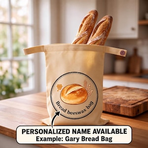 May include: A beige fabric bread bag with a circular graphic featuring a loaf of bread, wheat stalks, and a honeycomb pattern. The text reads "Bread beeswax bag". Two baguette loaves are inside the bag, which has a button closure on the side.