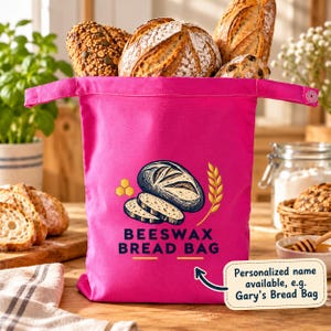 May include: A bright pink fabric bread bag featuring a graphic of a loaf of bread, honeycomb, and wheat stalks. The text reads BEESWAX BREAD BAG. A small attached tag includes an arrow pointing to the text: Personalized name available, e.g., Gary’s Bread Bag.