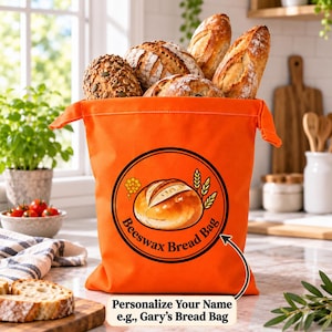 May include: A bright orange fabric bread bag filled with various loaves of crusty bread. The bag features a circular logo with an illustration of a round loaf, wheat stalks, and the text Beeswax Bread Bag. Below, text reads Personalize Your Name e.g., Gary's Bread Bag.