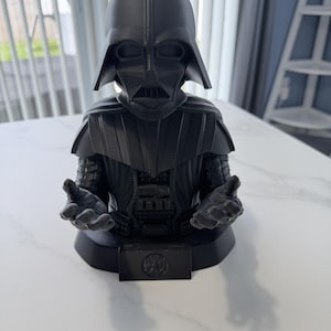 May include: A matte black 3D printed bust of Darth Vader featuring the character in a helmet and armor with outstretched hands. The base includes a small rectangular plaque with a circular Imperial crest emblem centered on the front surface.
