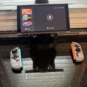 Könnte beinhalten: Eine schwarze Darth-Vader-Figur, die eine Nintendo Switch-Konsole mit einem dunklen Bildschirm hält, auf dem ein Startmenü angezeigt wird. Zwei abgenommene Joy-Con-Controller, einer mit hellblauen und einer mit orangen Akzenten, liegen davor.