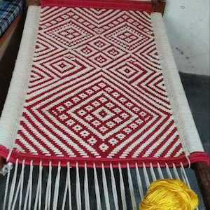 Könnte beinhalten: Ein rechteckiges Bett mit einer gewebten Oberfläche in einem geometrischen rot-weißen Muster. Der Rahmen besteht aus dunklem Holz mit geschnitzten Pfosten. Ein gelbes Garnknäuel liegt auf dem unvollendeten weißen Seilabschnitt am Fußende.